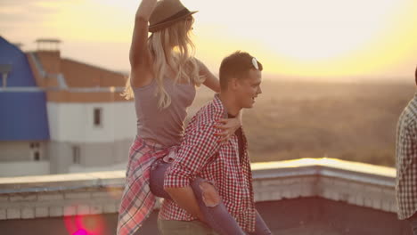 Loving-couple-moves-in-a-dance-at-a-party-with-friends.-A-girl-sits-on-her-boyfriend's-back-they-are-fooling-around-on-the-roof.